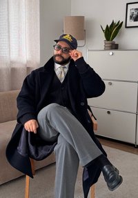 Homme en manteau noir, pantalon gris et casquette ajustant ses lunettes en étant assis jambes croisées sur une chaise en bois dans un salon moderne.