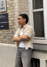 Beige nappipaita, lyhythihainen, valkoinen aluspaita, harmaat farkut ja mustat silmälasit. Seisoo tiiliseinän lähellä, jossa on kylttejä.