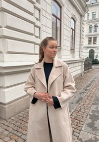 Hosszú, világos bézs gyapjúpulóver, gallérral, laza fazonnal, ejtett vállakkal és enyhén texturált anyaggal. Sötét pulóver felett viselve.