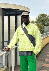 Νεον κίτρινο oversized μπουφάν με δύο μπροστινές τσέπες, γκρίζος διαγώνιος σάκος, γκρίζο πουκάμισο και πράσινα παντελόνια στίβου με λευκές ρίγες. Υφή υφάσματος.