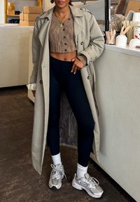 Beige trenchcoat over en brun kort jakke, svarte leggings og grå joggesko med hvite sokker. Innstillingen har en lys bakgrunn.