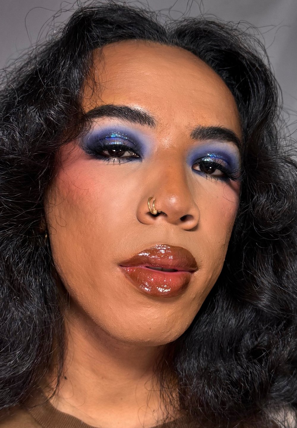 Person med mörkt lockigt hår, djärv blå glittrande ögonskugga, glansiga bruna läppar och guld näsringsar, iförd en brun topp.