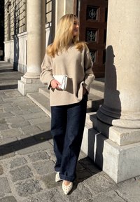 Bézs színű, kötött pulóver, széles ujjakkal, sötét farmernadrág és fehér, szegecses, hegyes orrú cipő. Egy kis fehér borítéktáskát tart. Városi környezetben.