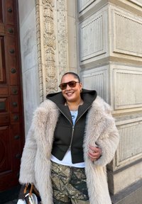 Long manteau en fausse fourrure crème porté sur une veste à capuche foncée, associé à un pantalon camouflage. Lunettes de soleil rondes et sac à main marron avec des accents blancs.