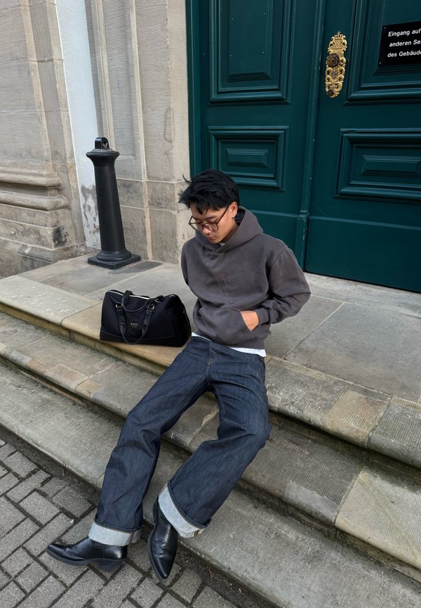 Jeune homme portant des lunettes et un sweat à capuche sombre assis sur des marches en pierre à côté d’un sac noir devant de grandes portes vertes avec une poignée dorée.