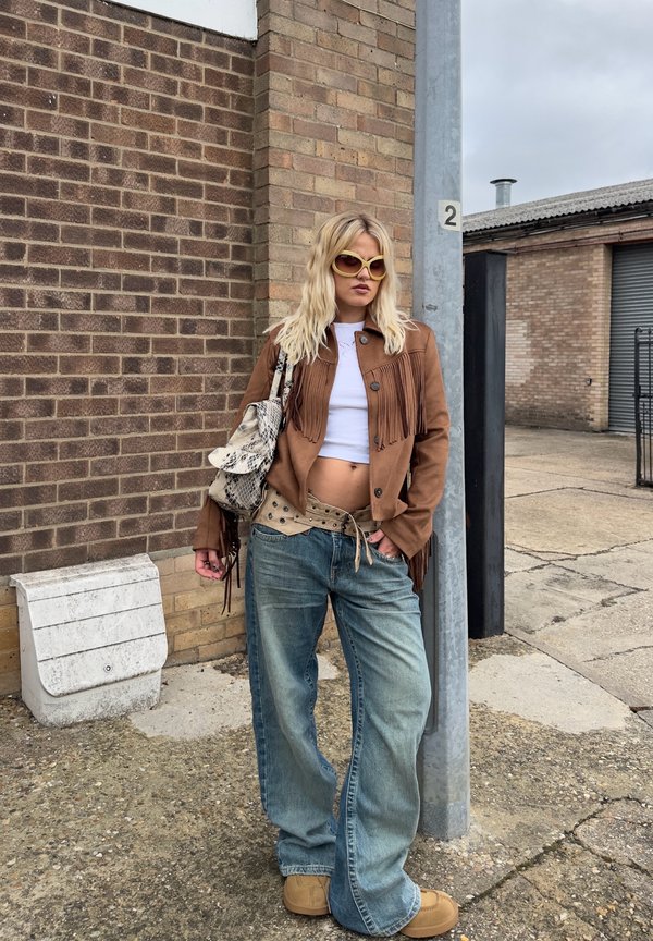 Junge Frau mit Sonnenbrille, brauner Fransjacke, weißem Kurztop, weitem Jeans und braunen Schuhen, die draußen vor einer Ziegelmauer steht.