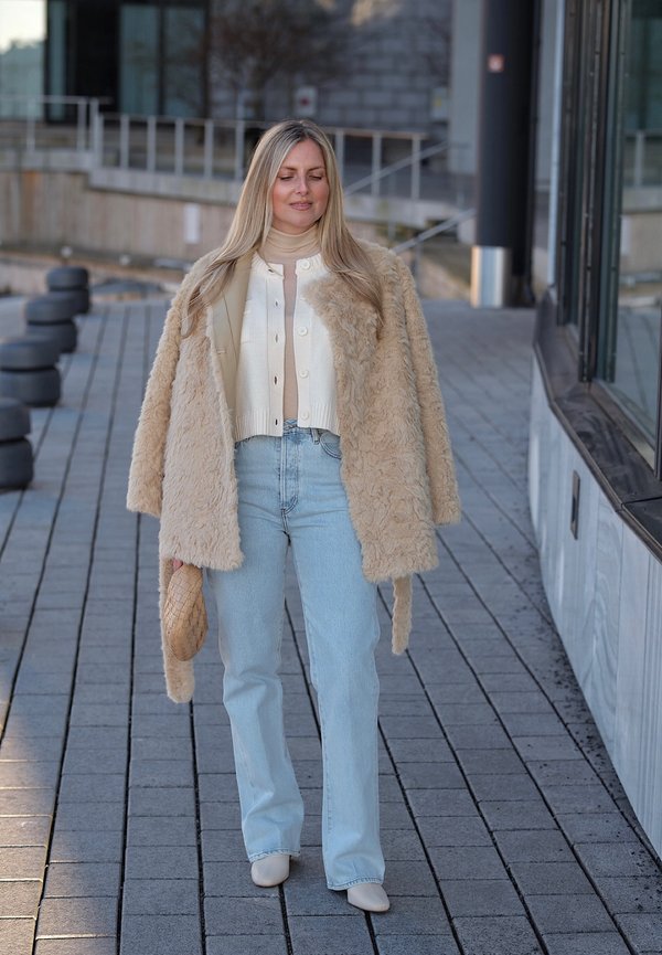 Mujer caminando al aire libre con un abrigo de piel sintética beige, cárdigan blanco, jeans azul claro, zapatos beige y llevando un bolso a juego.