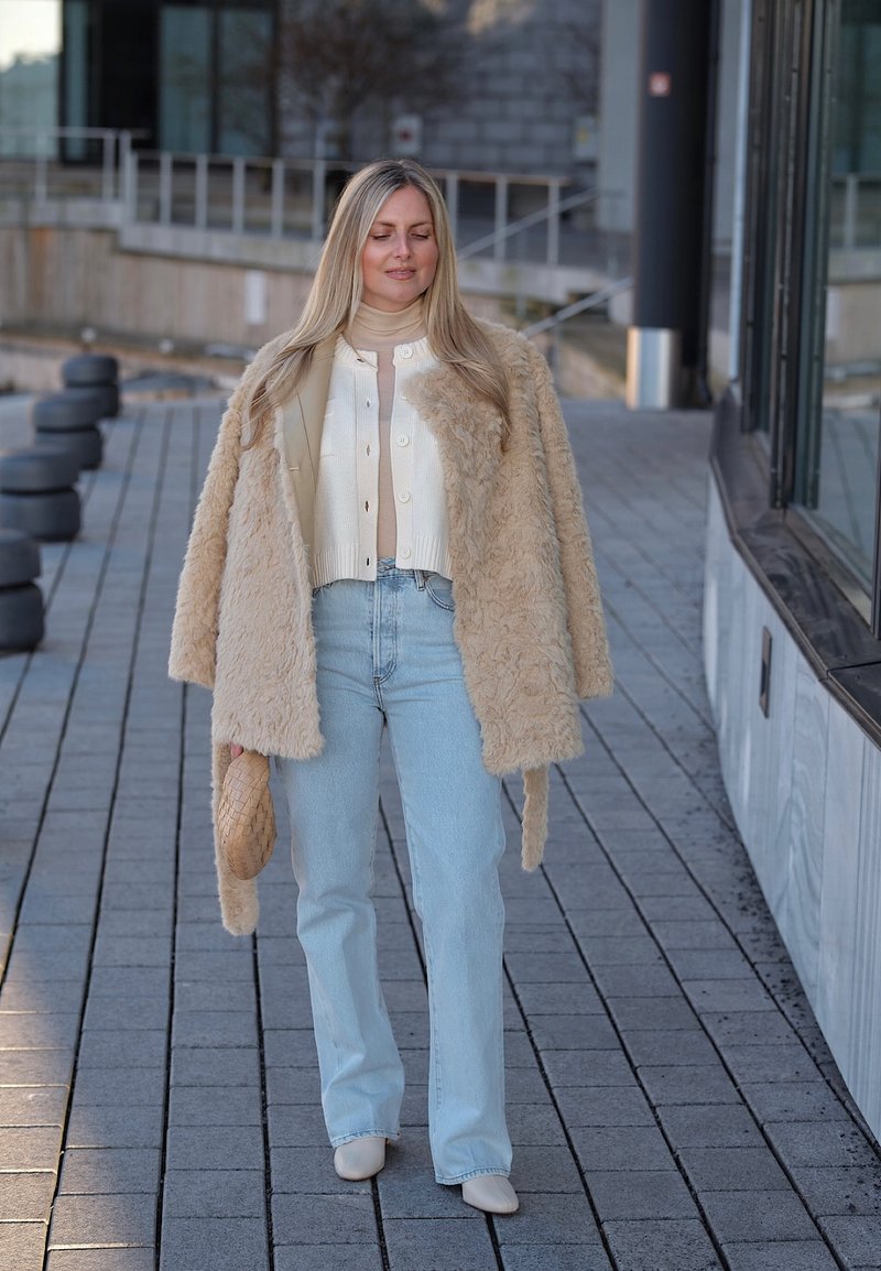 Frau geht draußen und trägt einen beigen Kunstpelzmantel, eine weiße Strickjacke, hellblaue Jeans, beigen Schuhe und hält eine passende Handtasche.