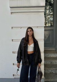 Μαύρο δερμάτινο oversized μπουφάν, λευκό crop top, ψηλόμεσα σκούρα τζιν, τσάντα με σχέδια και γυαλιά ηλίου. Ουδέτερο φόντο με πόρτα.