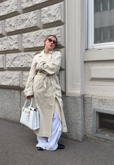 Persona con gabardina beige, pantalones anchos blancos, zapatos negros y gafas de sol apoyada en una pared de piedra texturizada sosteniendo un bolso blanco.