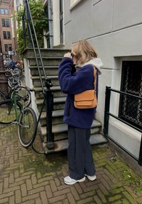 Veste polaire violette, capuche grise, pantalon gris clair et baskets grises ; porte un sac bandoulière marron. Des marches et des vélos en arrière-plan.