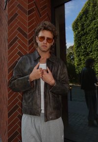 Brun skinnjakke med en subtil glans, glidelås, frontlommer, og grå joggebukser. Briller med oransjefarget glass. Murvegg i bakgrunnen.