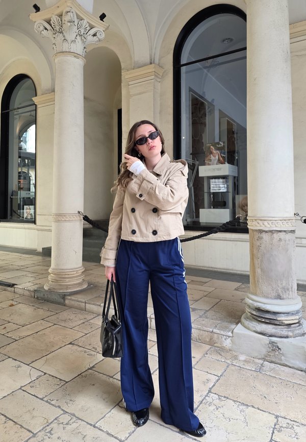 Beige double-breasted cropped jacket, navy wide-leg trousers with side stripes, black sunglasses, and a small black handbag. Stone columns in the background.