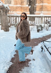 Veste en fausse fourrure marron, haut blanc, jeans évasés bleu clair et bottes à talons marron. Fond enneigé avec un chemin en briques et un banc.