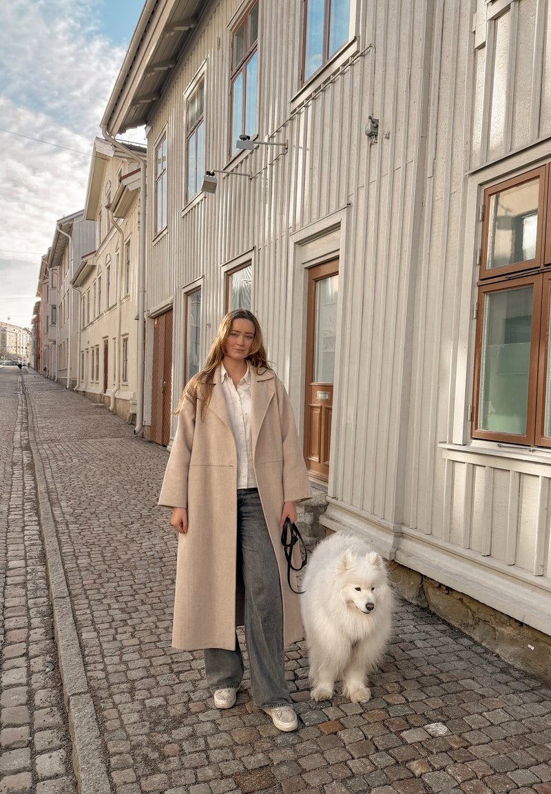 Femme en manteau beige promenant un grand chien blanc et duveteux dans une rue pavée bordée de bâtiments en bois clair au coucher du soleil.