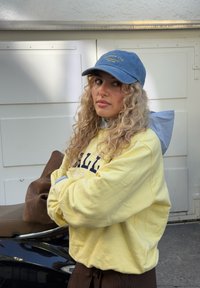 Giovane donna con capelli biondi ricci che indossa un berretto blu e una felpa gialla, in piedi con le braccia incrociate vicino a uno scooter scuro all'aperto.