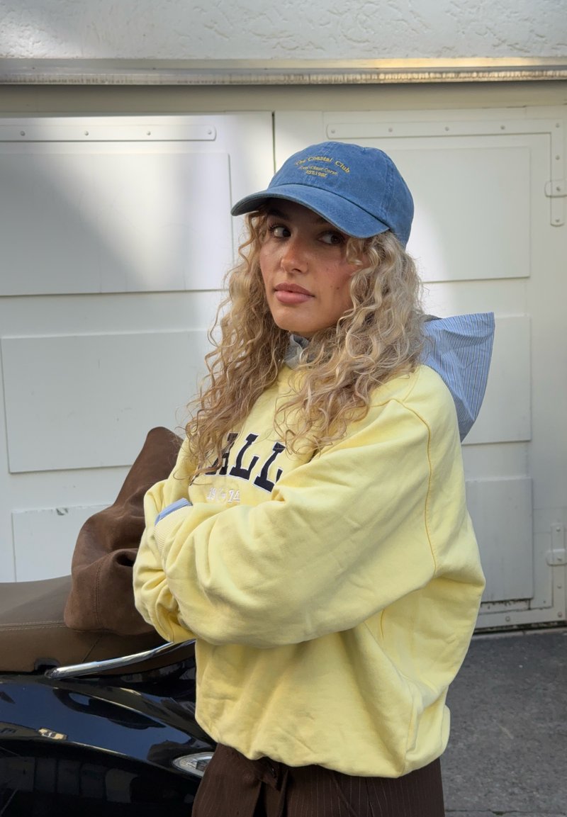 Jeune femme aux cheveux blonds bouclés portant une casquette bleue et un sweat-shirt jaune, debout les bras croisés près d'un scooter sombre en plein air.