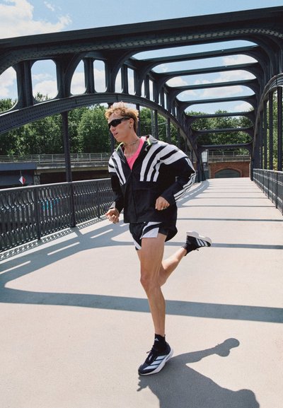 Chaqueta atlética en blanco y negro con rayas, camisa rosa, pantalones cortos negros y zapatillas. Corriendo sobre un puente con un diseño de arco de metal.