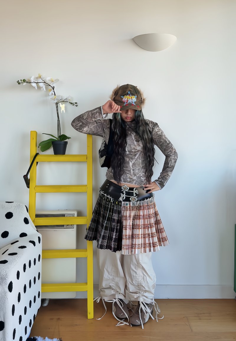 Camouflage lange mouwen top, geruite rok in tinten van zwart, bruin en oranje, leren riem met zilveren accenten, ruime witte broek en een pet.