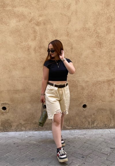 Top corto negro, pantalones cortos de denim beige con bordes deshilachados, zapatillas negras y rojas, y un bolso verde. Unas gafas de sol y joyas completan el look.