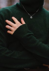 Pullover a collo alto verde scuro con polsini a coste, presentante una collana d'argento con un pendente quadrato e un piccolo dettaglio in pietra.