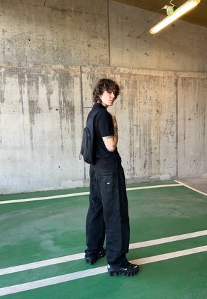 Camiseta negra, pantalones cargo negros holgados y zapatillas negras. Mochila negra pequeña llevada en la espalda. Pared de concreto y suelo de estacionamiento verde visibles.