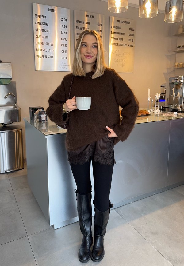 Femme en pull marron et bottes noires tenant une tasse blanche, debout dans un café moderne avec un comptoir en métal et des panneaux de menu derrière elle.