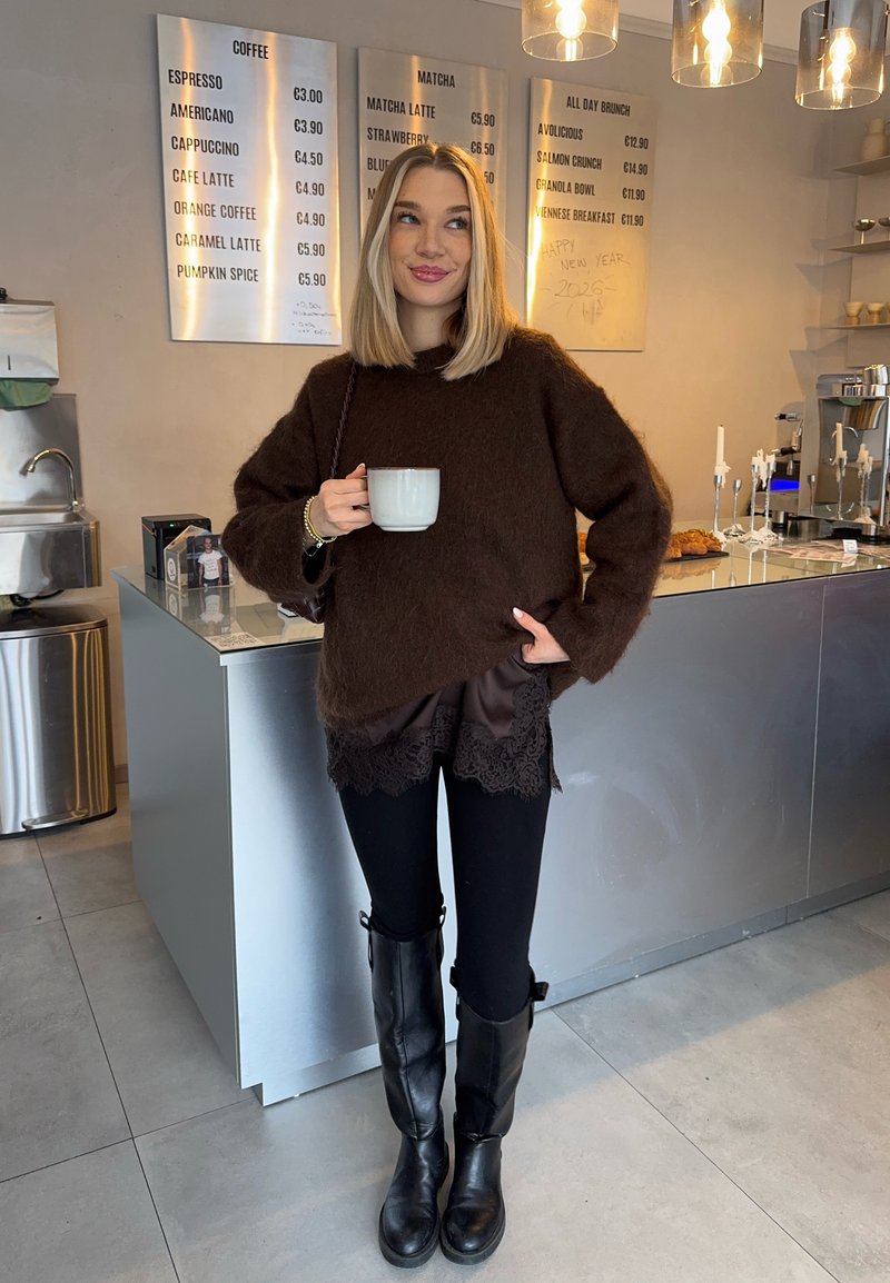 Femme en pull brun et bottes noires tenant une tasse blanche, debout dans un café moderne avec un comptoir en métal et des panneaux de menu derrière elle.