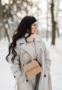 Femme en manteau beige et col roulé, portant des lunettes de soleil et un bandeau, tenant un sac à main beige en plein air dans une forêt enneigée.