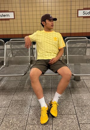 Camisa a rayas amarillas, pantalones cortos caquis, calcetines blancos y zapatos amarillos. Sentado en bancos de metal en una estación de metro con azulejos.