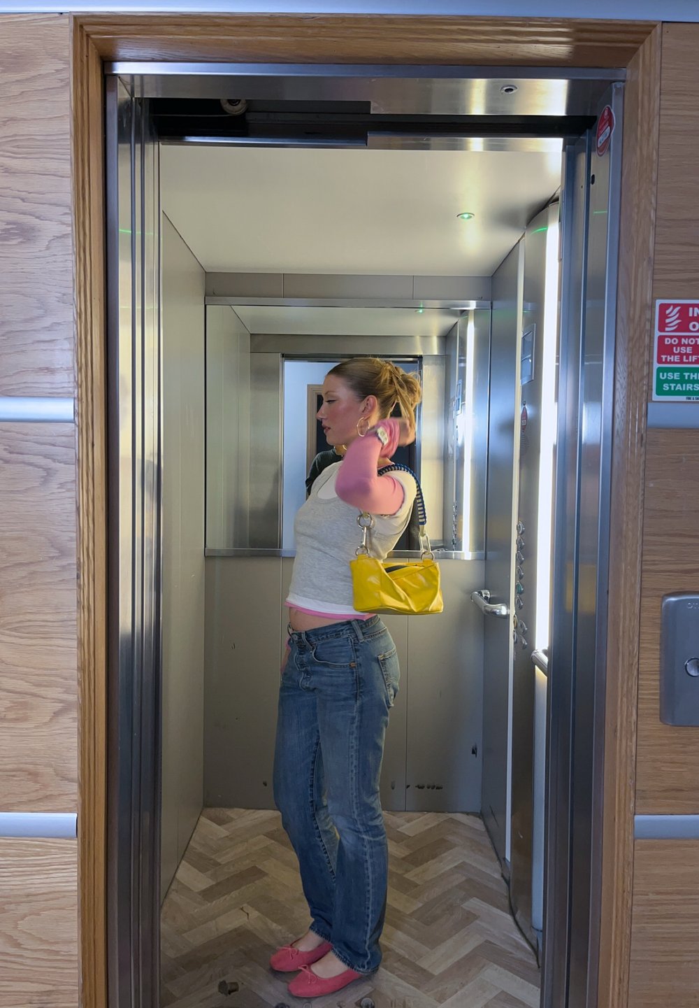 Jeune femme avec un sac à main jaune ajustant ses cheveux à l'intérieur d'un ascenseur moderne avec des panneaux en bois à l'extérieur de la porte.