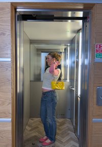 Jeune femme avec un sac à main jaune ajustant ses cheveux à l'intérieur d'un ascenseur moderne avec des panneaux en bois à l'extérieur de la porte.