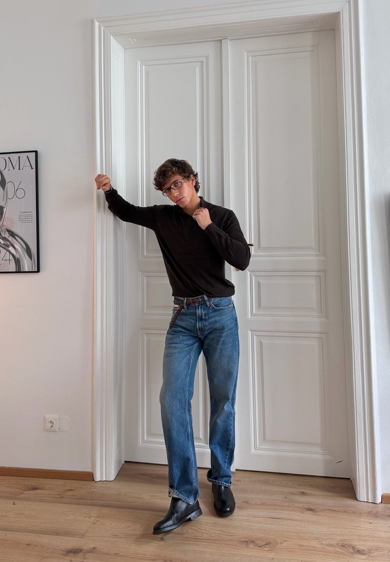 Schwarzes Langarm-Polohemd, hellblaue gerade Jeans, schwarze Knöchel-Stiefel; steht neben weißen Doppeltüren, Holzboden, minimalistische Dekoration.