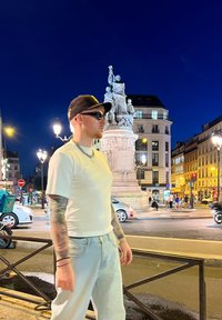 Una persona in maglietta bianca e jeans azzurri chiari si trova vicino a un monumento di pietra. Sono visibili un cappellino nero, occhiali da sole e tatuaggi. Ambientazione notturna.