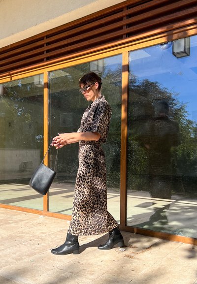 Vestido midi con estampado de leopardo, mangas cortas, botas negras hasta el tobillo y un pequeño bolso negro, todo en un fondo de pared de cristal.