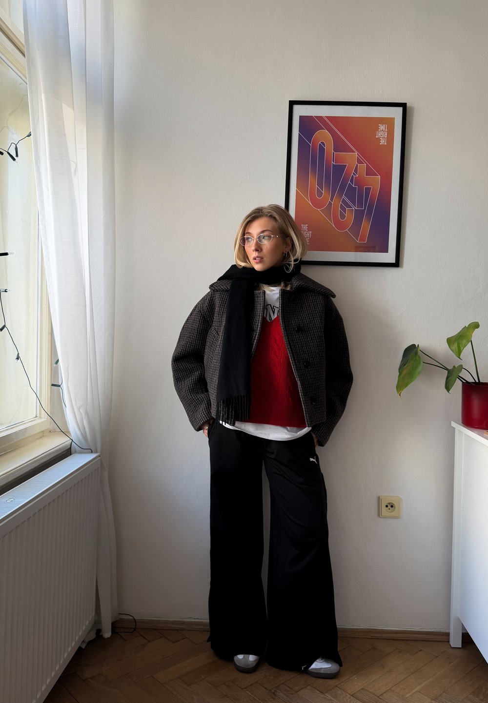 Veste à carreaux noirs et gris, pull en laine rouge, t-shirt blanc, pantalon noir à jambes larges, chaussures argentées, lunettes translucides, environnement intérieur avec plante et œuvre d'art.