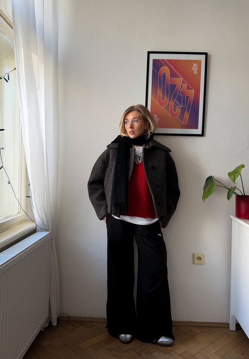 Black and gray checked jacket, red knitted sweater, white t-shirt, black wide-leg pants, silver shoes, clear glasses, indoor setting with plant and art.