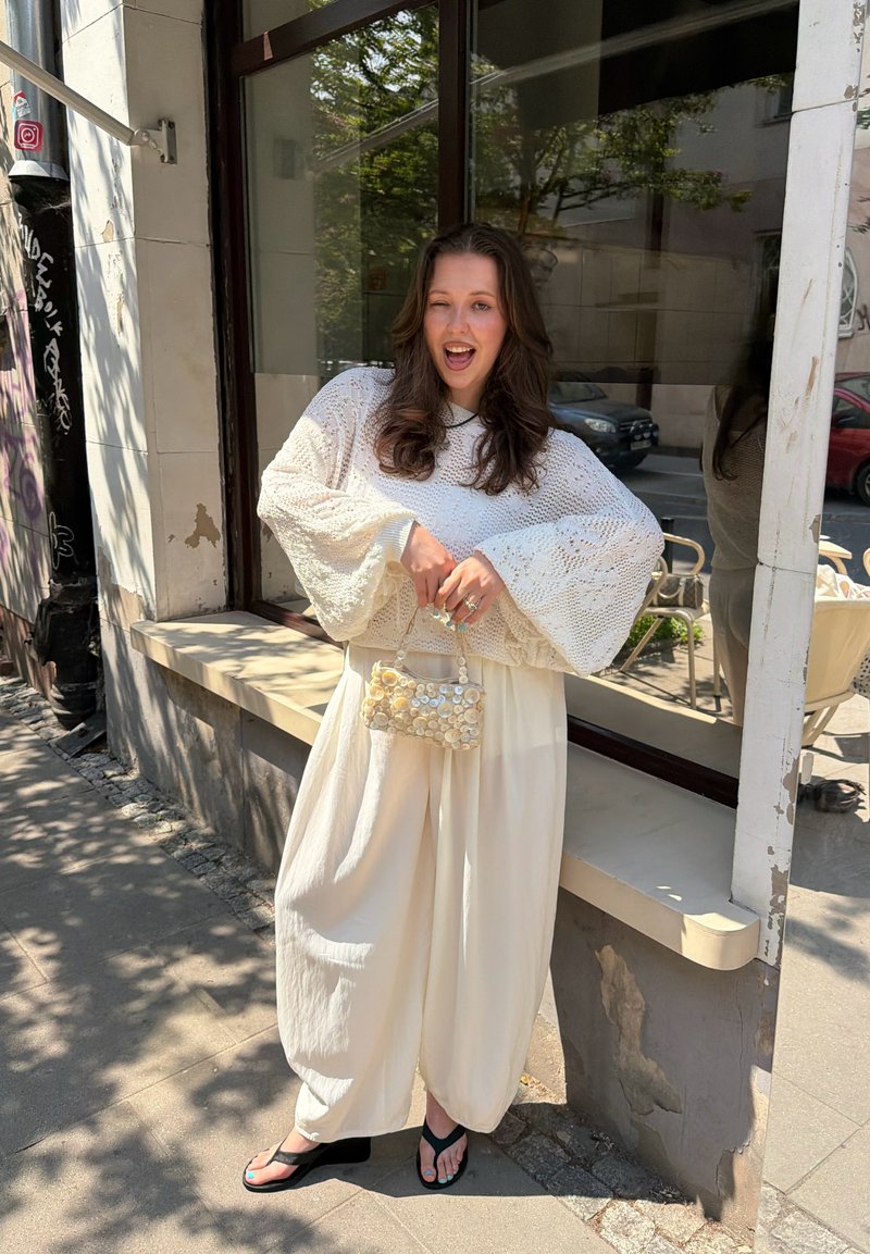 Femme en pantalon blanc ample et pull en maille tenant un petit sac perlé, debout sur le trottoir près d'une fenêtre de bâtiment par une journée ensoleillée.