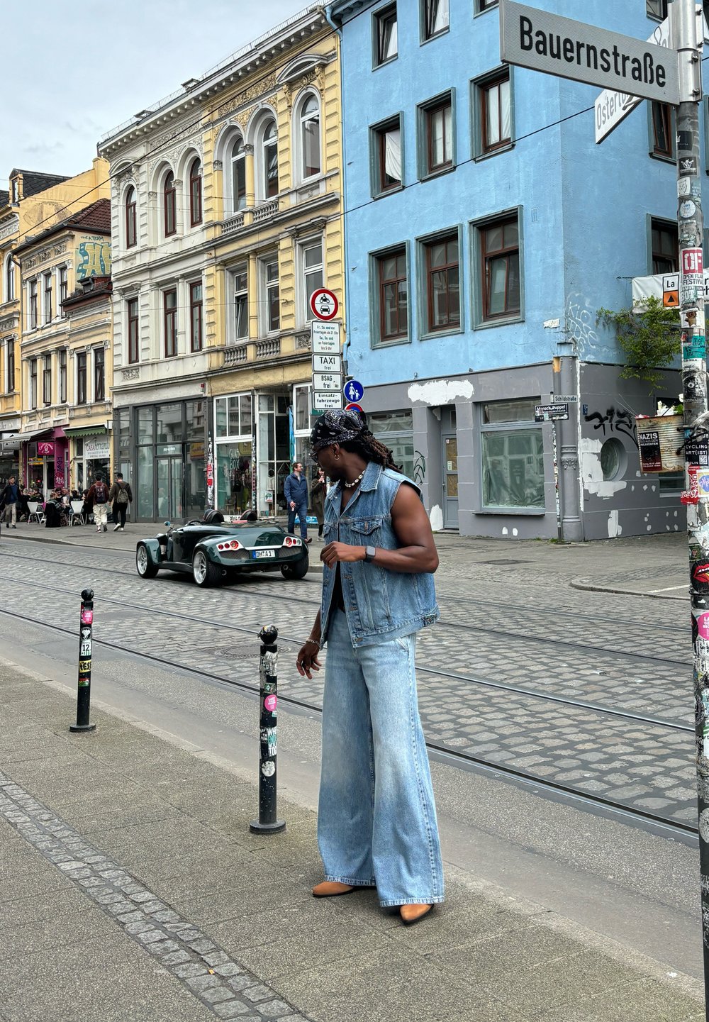Chaleco de mezclilla y vaqueros de pierna ancha en un lavado claro, complementados con tacones marrones; calle urbana con edificios y un coche vintage en el fondo.