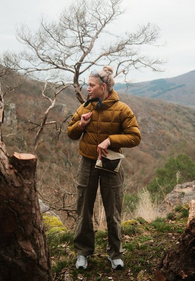 Chaqueta de plumas marrón, pantalones cargo olivos, sosteniendo un pequeño bolso blanco y negro. Entorno al aire libre con árboles desnudos y un paisaje rocoso.