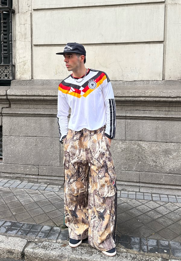 Ung mann iført en Tyskland fotballdrakt, vide kamuflasjebukser med lommer, svart caps og joggesko, stående mot en steinvegg på fortauet.