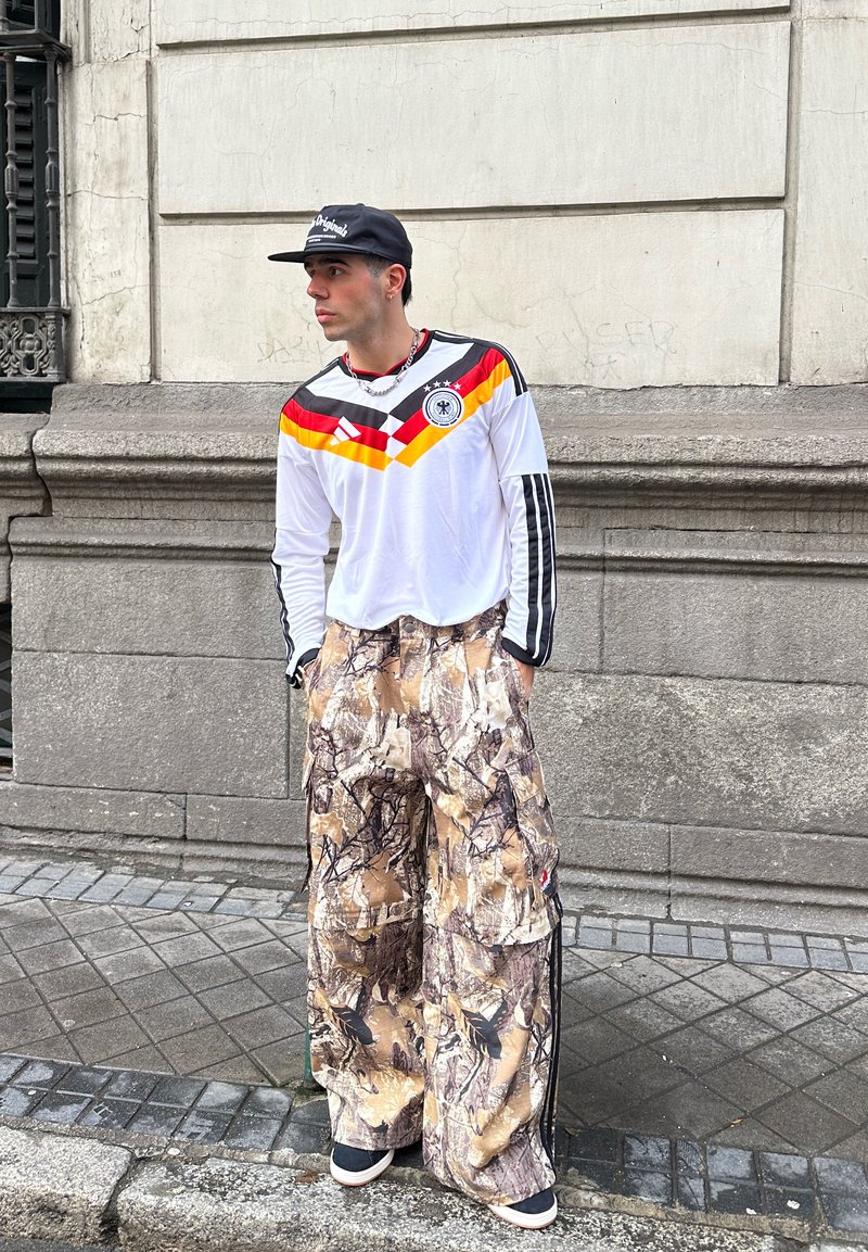 Νεαρός άνδρας με φανέλα ποδοσφαίρου της Γερμανίας, φαρδιά παντελόνια καμουφλάζ cargo, μαύρο καπέλο και αθλητικά παπούτσια, στέκεται μπροστά σε έναν πέτρινο τοίχο σε πεζοδρόμιο.