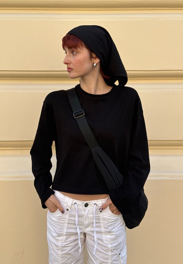Junge Person mit roten Haaren, die ein schwarzes Kopftuch, ein schwarzes Langarmshirt, weiße Hose und eine schwarze Umhängetasche trägt und nach links schaut.