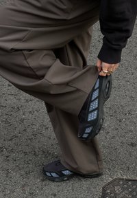 Sorte atletikskøjter med grå accenter, der har et let design og tekstureret sål, båret med løstsiddende brune bukser på en asfaltoverflade.