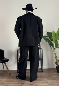 Completo nero con giacca oversize e pantaloni a gamba larga, abbinato a un cappello nero a tesa larga. Tessuto morbido, design minimale senza fantasie.