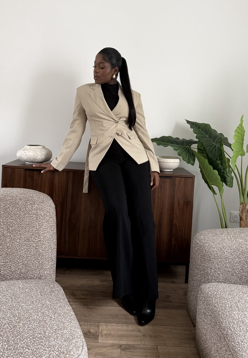 Femme en blazer beige et pantalon noir appuyée sur un buffet en bois avec des bols décoratifs et une grande plante verte dans un salon moderne.