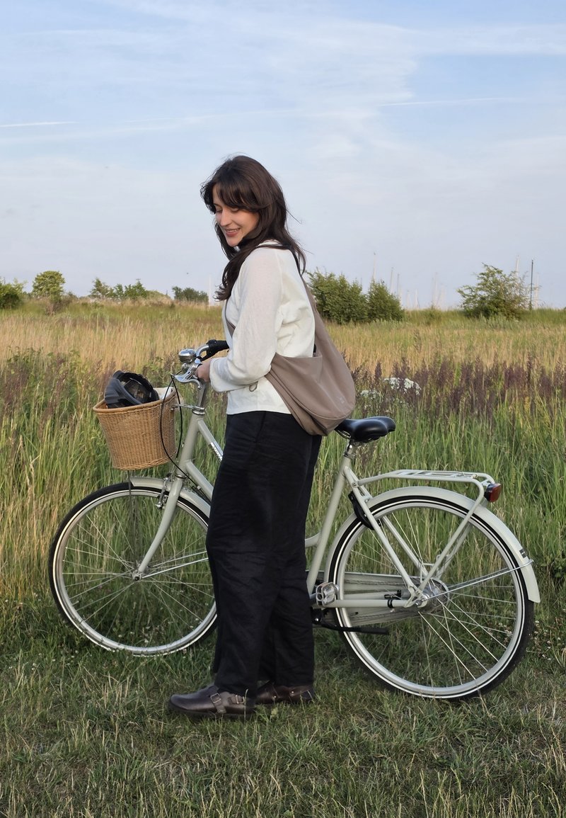 Woman standing outdoors holding a bicycle with a wicker basket containing a helmet in a grassy field under a cloudy sky.