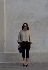 Beżowa, krótka peleryna z wysokim kołnierzem, czarne legginsy i czarne, spiczaste buty. Trzyma teksturowaną czarną kopertówkę. Jednolity tle.