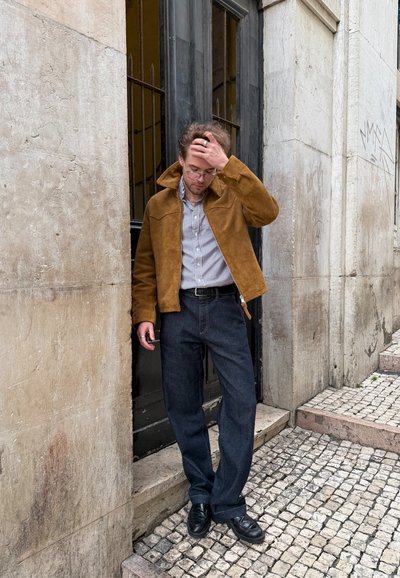 Joven con chaqueta marrón y camisa de rayas que se ajusta el cabello mientras está de pie en escalones de piedra frente a un edificio con una puerta alta negra y pavimento de adoquines.
