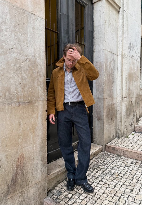 Młody mężczyzna w brązowej kurtce i pasiastym t-shircie poprawia włosy stojąc na kamiennych schodach przed budynkiem z wysokimi czarnymi drzwiami i brukowanym chodnikiem.